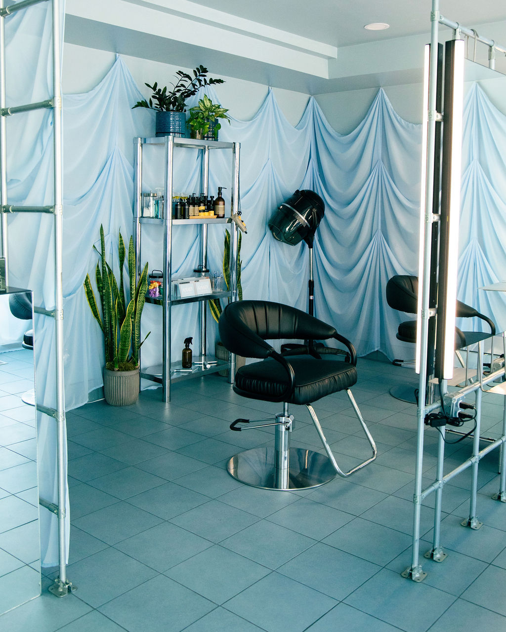 Light-blue modern hair salon interior with black styling chairs, hooded dryer, chrome shelving stocked with hair products and potted plants, draped fabric walls and tiled floor