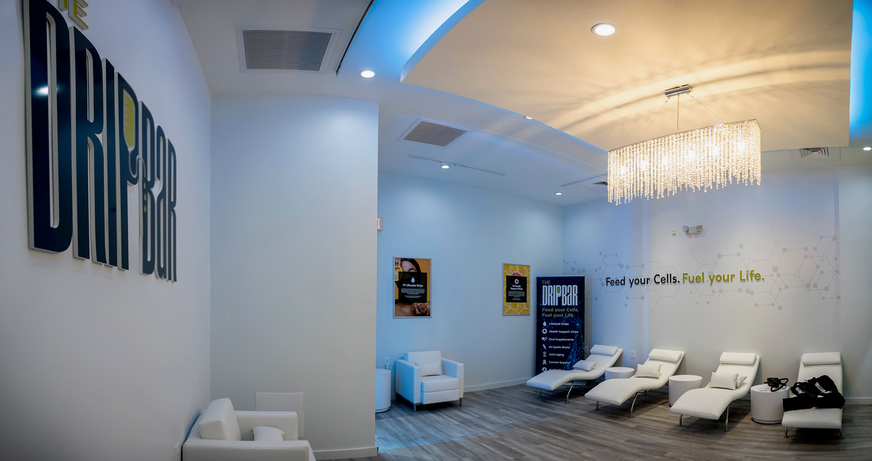 Modern wellness clinic IV therapy lounge with white reclining treatment chairs, a crystal chandelier, blue-accent ceiling lighting, hardwood floors, and minimalist wall graphics