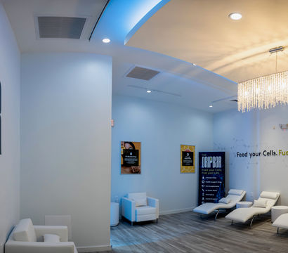 Modern wellness clinic IV therapy lounge with white reclining treatment chairs, a crystal chandelier, blue-accent ceiling lighting, hardwood floors, and minimalist wall graphics