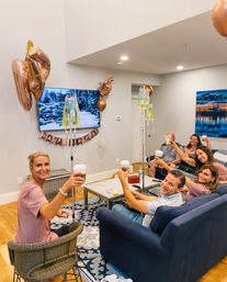 Friends toasting with coffee in a cozy living room birthday party, rose-gold balloons, "Happy Birthday" banner and IV drip poles nearby.