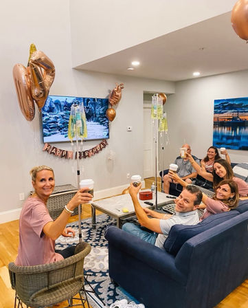 Friends toasting with coffee in a cozy living room birthday party, rose-gold balloons, "Happy Birthday" banner and IV drip poles nearby.