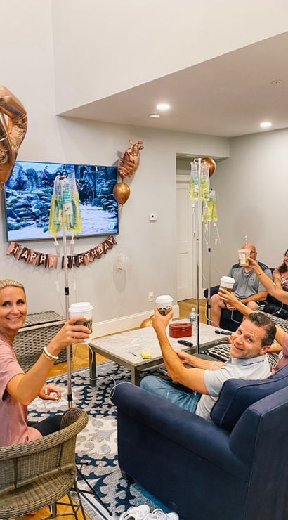 Friends toasting with coffee in a cozy living room birthday party, rose-gold balloons, "Happy Birthday" banner and IV drip poles nearby.