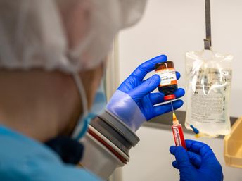 Gloved healthcare worker in a hospital clinical setting draws medication from an amber vial into a syringe, with a hanging IV saline bag visible in the background.