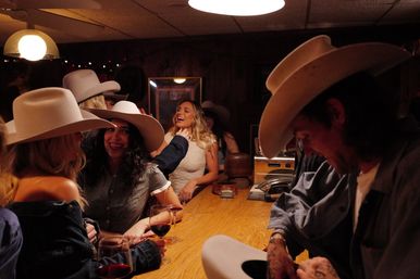 Group of friends wearing cowboy hats laughing at a rustic wood-paneled saloon bar, toasting with red wine under warm dim lights.