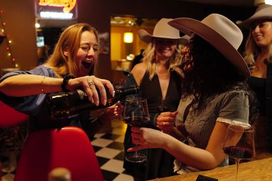 Women in cowboy hats laughing and sharing red wine at a cozy western-style bar during a night out