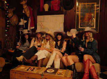 Six women in cowboy hats and boots laughing and holding wine glasses on a rustic couch in an Austin, Texas–style western lounge with mounted deer heads, a wall map and vintage poster