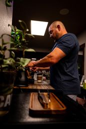 Barista preparing pour-over coffee at a cozy neighborhood café counter, paper to-go cup and potted plants in the foreground under warm overhead lighting.