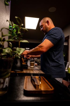 Barista preparing pour-over coffee at a cozy neighborhood café counter, paper to-go cup and potted plants in the foreground under warm overhead lighting.