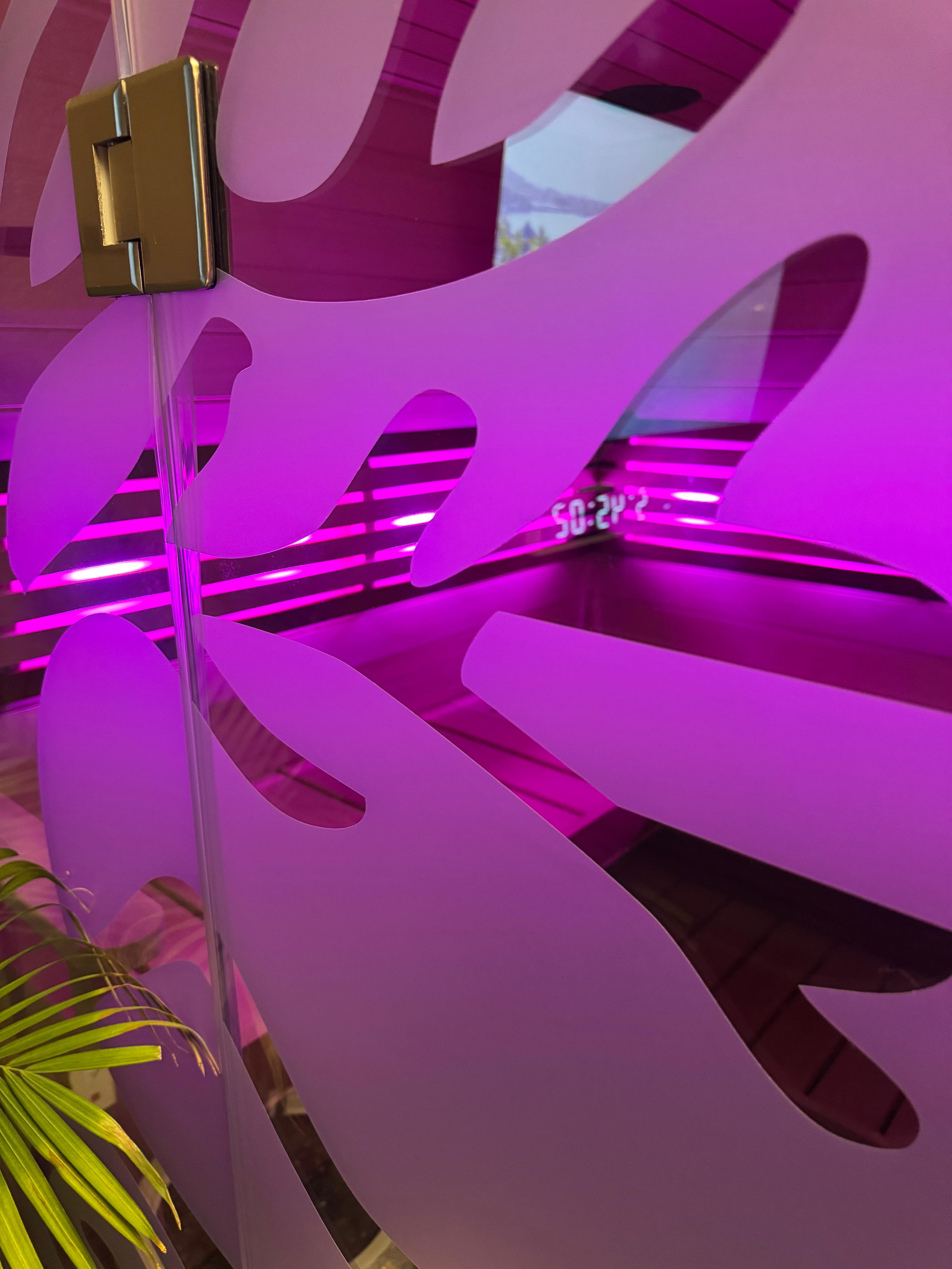 Frosted glass partition with oversized leaf cutouts revealing vibrant magenta LED backlighting and a faint digital clock reflection; tropical plant frond at lower-left, modern indoor decor.