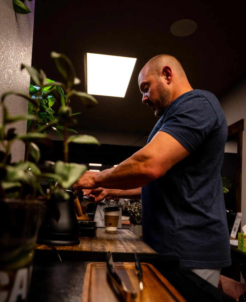 Barista preparing espresso at a wooden counter in a cozy coffee shop interior, potted plants in the foreground and a takeaway cup waiting under the portafilter.