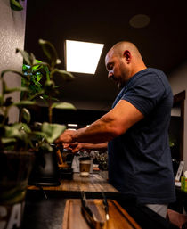 Barista preparing espresso at a wooden counter in a cozy coffee shop interior, potted plants in the foreground and a takeaway cup waiting under the portafilter.