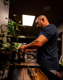 Person making espresso at a cafe counter, tamping coffee and pouring into a to-go cup under a bright overhead light with potted plants blurred in the foreground