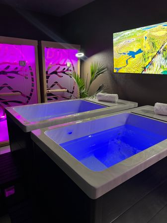Modern indoor spa relaxation room with two glowing blue float therapy tubs, purple backlit cubicles, rolled towels, tropical plant and wall landscape art.