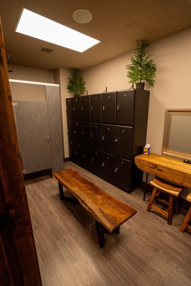 Cozy modern locker room with live-edge wooden bench, stacked black lockers, small wood vanity and stools, potted greenery and gray wood-look flooring under warm overhead lighting