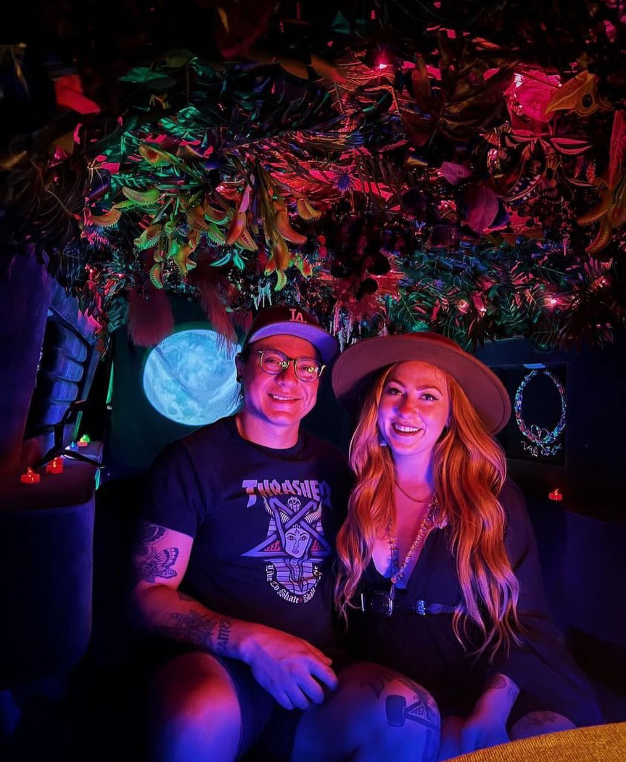 Smiling couple in a neon-lit indoor tropical installation — hanging foliage and colorful purple-pink-blue lights, moon projection behind them; woman in a wide-brim hat and long hair, man with glasses and tattooed arms, playful date-night vibe.
