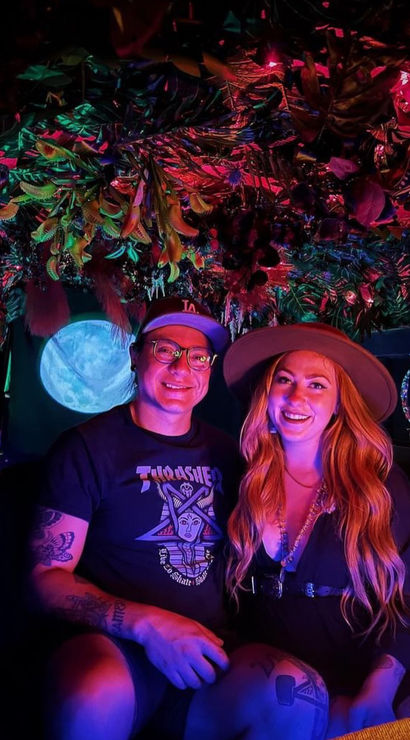 Smiling couple in a neon-lit indoor tropical installation — hanging foliage and colorful purple-pink-blue lights, moon projection behind them; woman in a wide-brim hat and long hair, man with glasses and tattooed arms, playful date-night vibe.