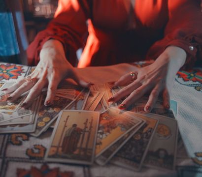 Candlelit tarot reading — fortune-teller's hands in a red blouse fanning tarot cards across a colorful embroidered tablecloth, with a lit candle and hourglass nearby.
