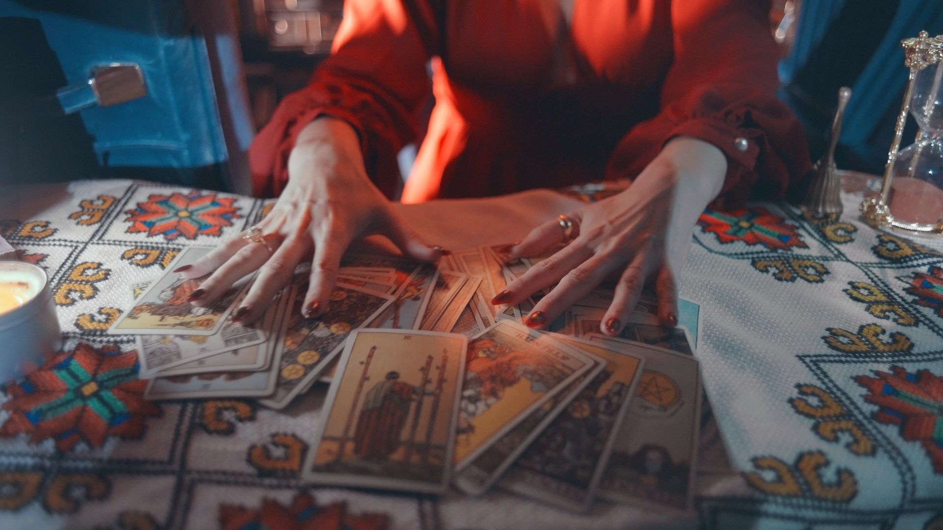 Candlelit tarot reading — fortune-teller's hands in a red blouse fanning tarot cards across a colorful embroidered tablecloth, with a lit candle and hourglass nearby.