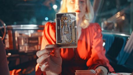 Person in a red blouse holding a High Priestess tarot card to the camera inside a vintage car at night, mystic vibe with bokeh city lights and retro dashboard visible.