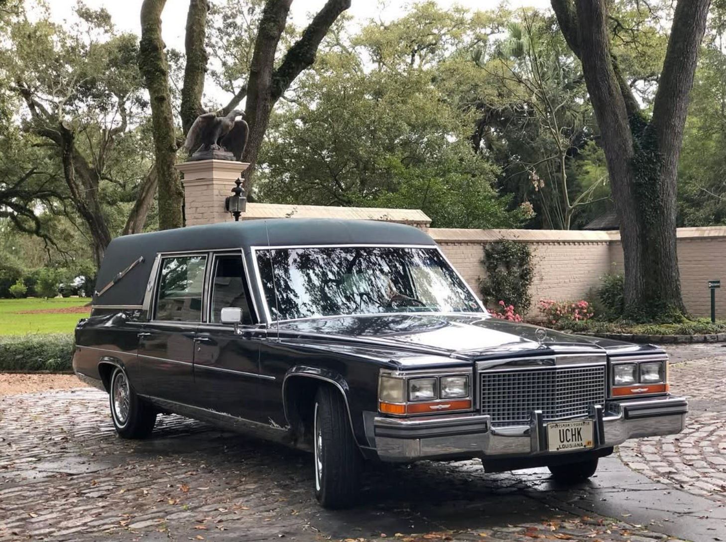 Vintage black Cadillac hearse parked on a cobblestone driveway beneath live oak trees, with a brick garden wall and eagle statue in a lush Louisiana setting.