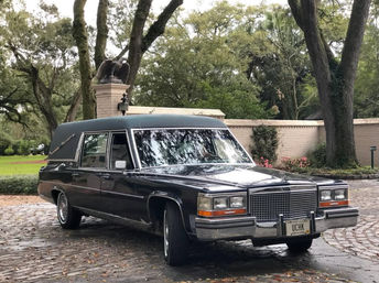 Vintage black Cadillac hearse parked on a cobblestone driveway beneath live oak trees, with a brick garden wall and eagle statue in a lush Louisiana setting.
