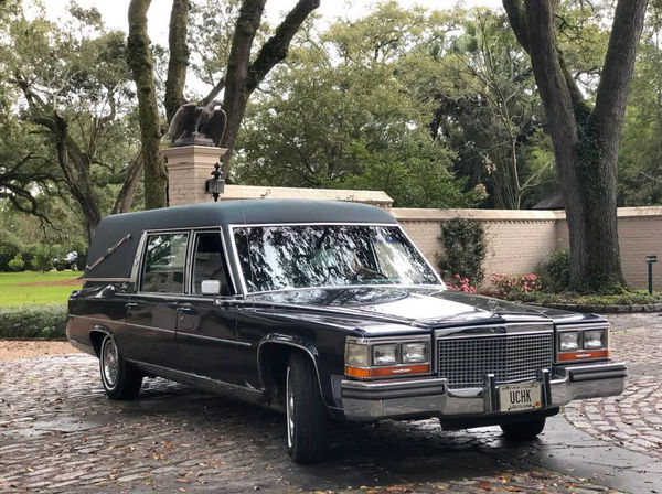 Vintage black Cadillac hearse parked on a cobblestone driveway beneath live oak trees, with a brick garden wall and eagle statue in a lush Louisiana setting.