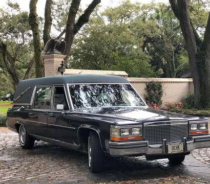 Vintage black Cadillac hearse parked on a cobblestone driveway beneath live oak trees, with a brick garden wall and eagle statue in a lush Louisiana setting.