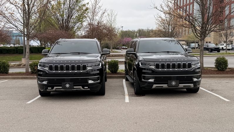 Two black Wagoneer SUVs parked side-by-side, front view in a suburban office parking lot with early-spring trees, landscaped shrubs and an overcast sky.