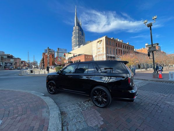 Sunny downtown Nashville street scene with a shiny black SUV in the foreground, brick storefronts and a dramatic spired skyscraper against a bright blue sky.
