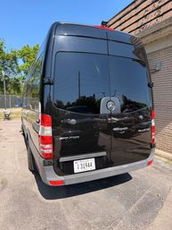 Rear view of a glossy black Mercedes Sprinter cargo van parked by a brick building in a sunny parking lot, blue sky and trees reflected in the tinted rear doors.