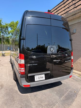 Rear view of a glossy black Mercedes Sprinter cargo van parked by a brick building in a sunny parking lot, blue sky and trees reflected in the tinted rear doors.