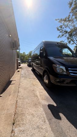 Sleek black passenger van parked beside a brick building in a sunny urban alley, blue sky overhead, trees in the background and long sidewalk shadows.