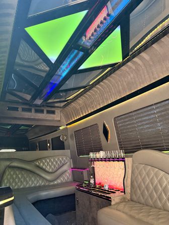 Luxury party limousine interior with quilted gray leather seats, LED-lit mirrored ceiling panels in green and blue, built-in champagne glass rack and pink ambient accent lighting.
