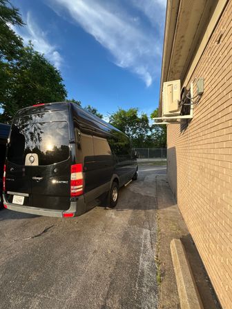 Shiny black passenger van parked beside a beige brick building with a wall-mounted HVAC unit, sun reflecting on the van, wispy blue sky and trees in a suburban parking lot.