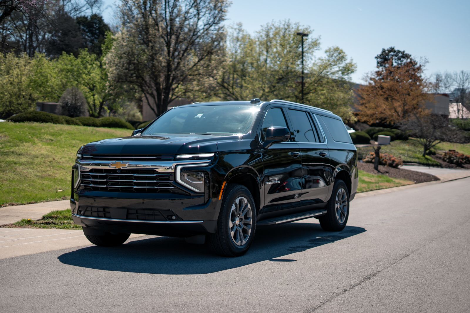 Shiny black Chevrolet Suburban SUV parked curbside in a sunny suburban neighborhood with green lawns and trees