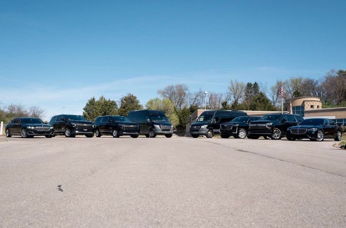 Row of black luxury sedans, SUVs and passenger vans lined up in a suburban parking lot outside an office building with an American flag under a clear blue sky
