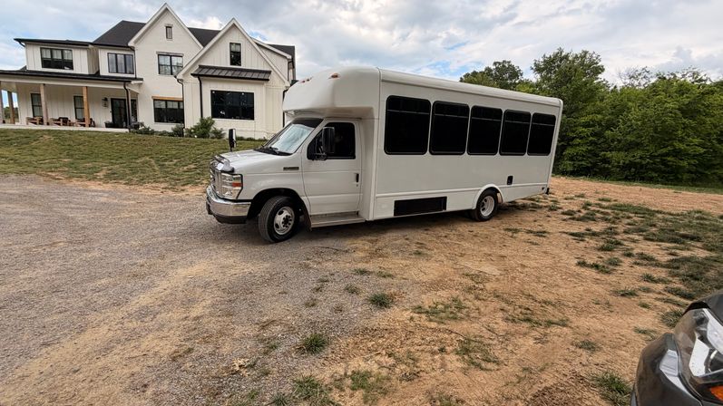 White passenger shuttle bus parked on a gravel driveway beside a modern white farmhouse with porch and trees under a cloudy sky.