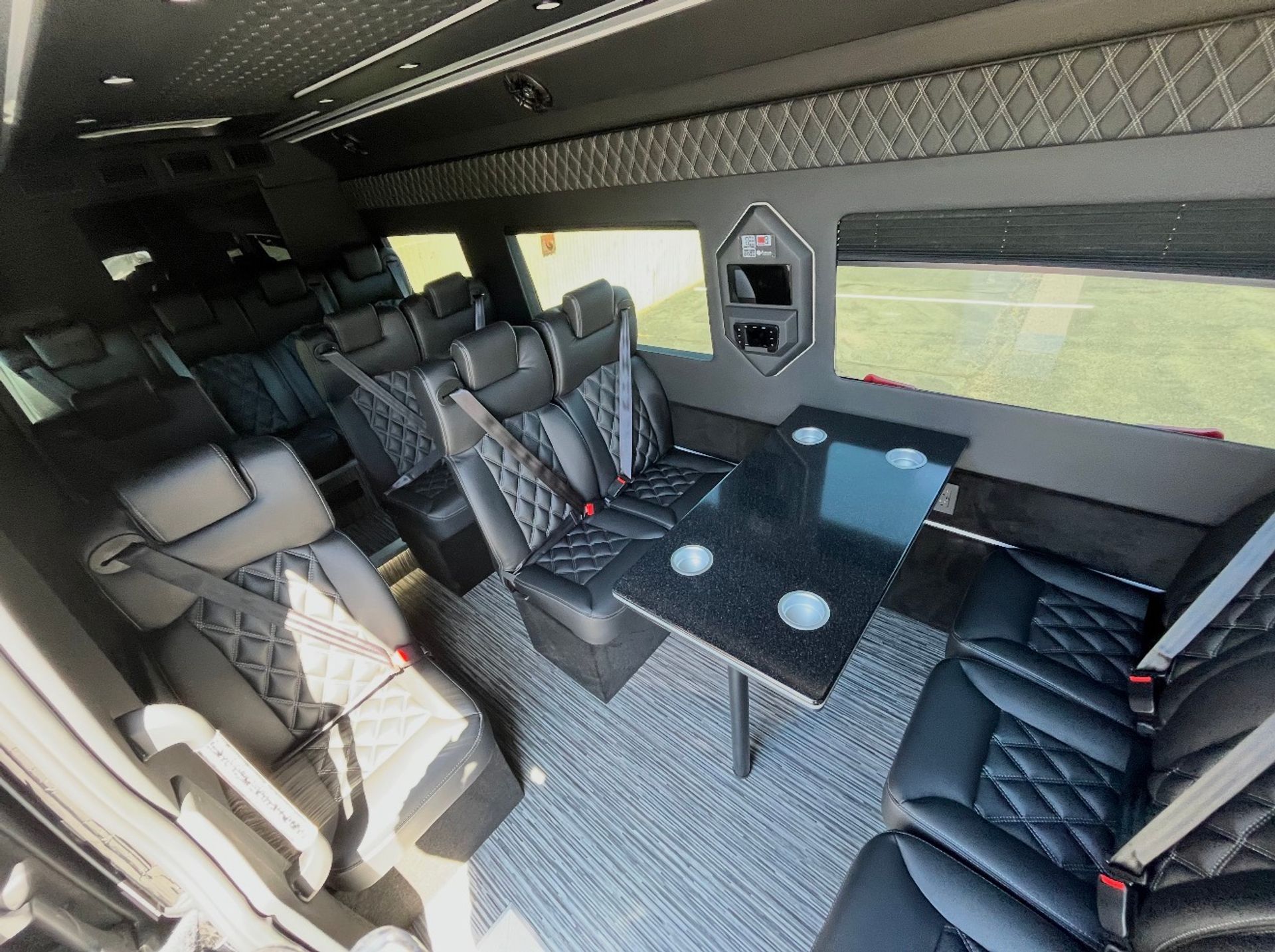 Luxury passenger van interior with black quilted leather seats, seatbelts, a glossy table with four cup holders and large side windows overlooking green lawn — executive shuttle style.