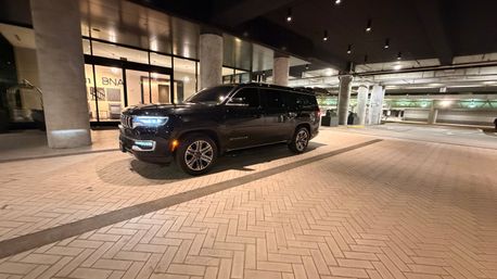 Black luxury SUV parked under a lit covered drop-off at night, headlights glowing beside concrete columns and patterned brick pavement in front of a building entrance.