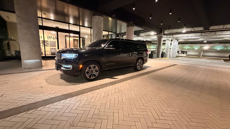 Black luxury SUV parked under a lit covered drop-off at night, headlights glowing beside concrete columns and patterned brick pavement in front of a building entrance.