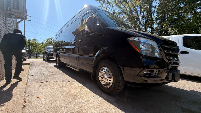 Shiny black Mercedes Sprinter 3500 cargo van parked on a sunlit driveway with tree and sky reflections, other vehicles nearby and a person bending by a brick wall.