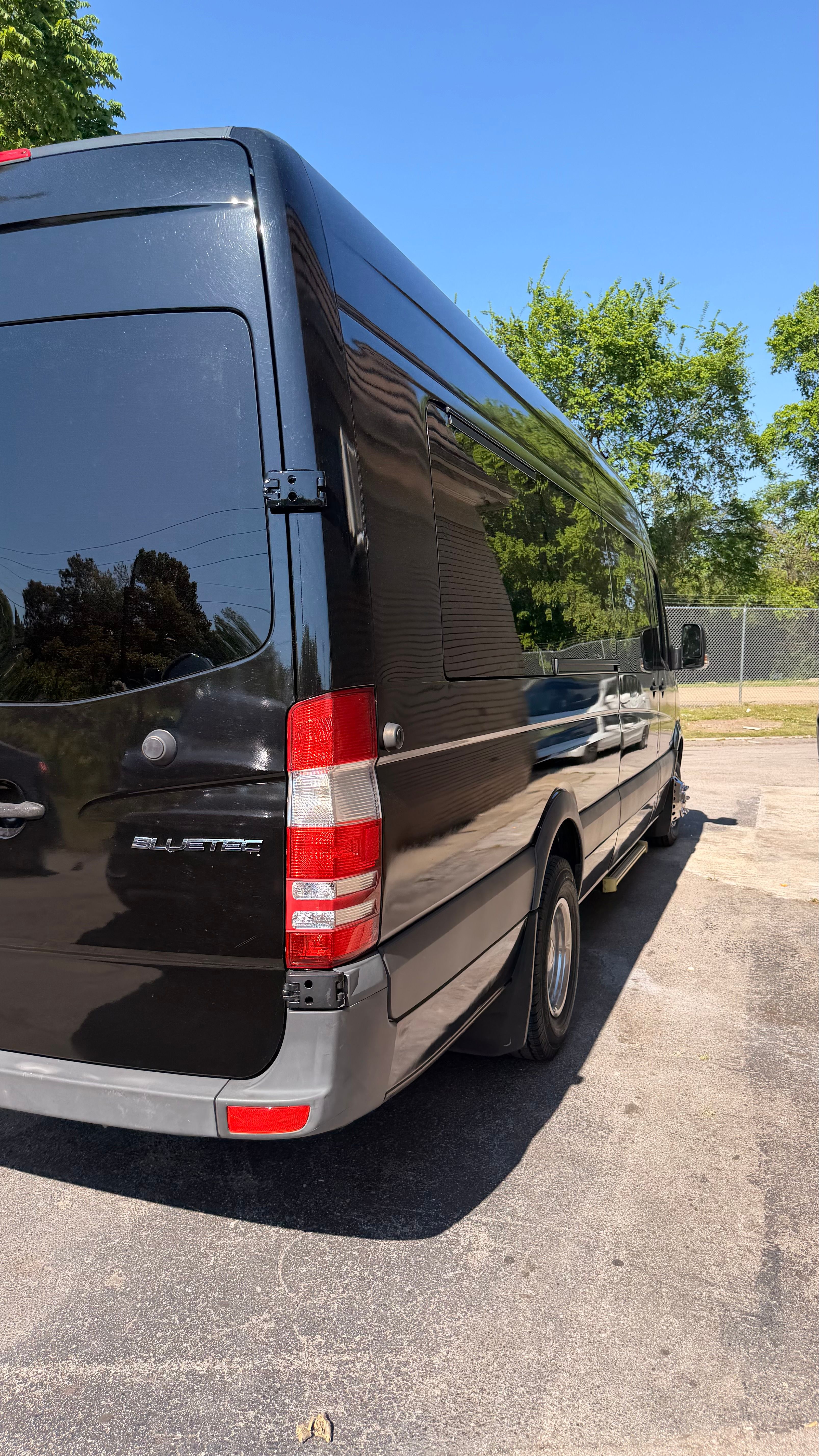 Rear three-quarter view of a black cargo van parked in a sunny suburban lot, reflective side panels mirroring trees and a house under a clear blue sky