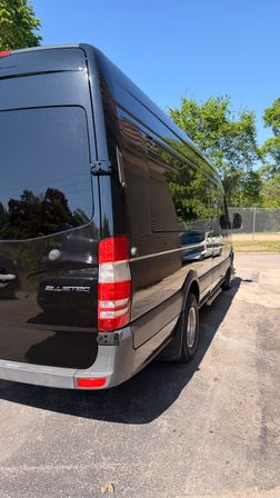 Rear three-quarter view of a black cargo van parked in a sunny suburban lot, reflective side panels mirroring trees and a house under a clear blue sky