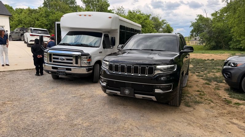 Black Wagoneer SUV and white passenger shuttle van parked side-by-side on a gravel driveway at a rural property, surrounded by other SUVs, a few people, green trees and a cloudy sky
