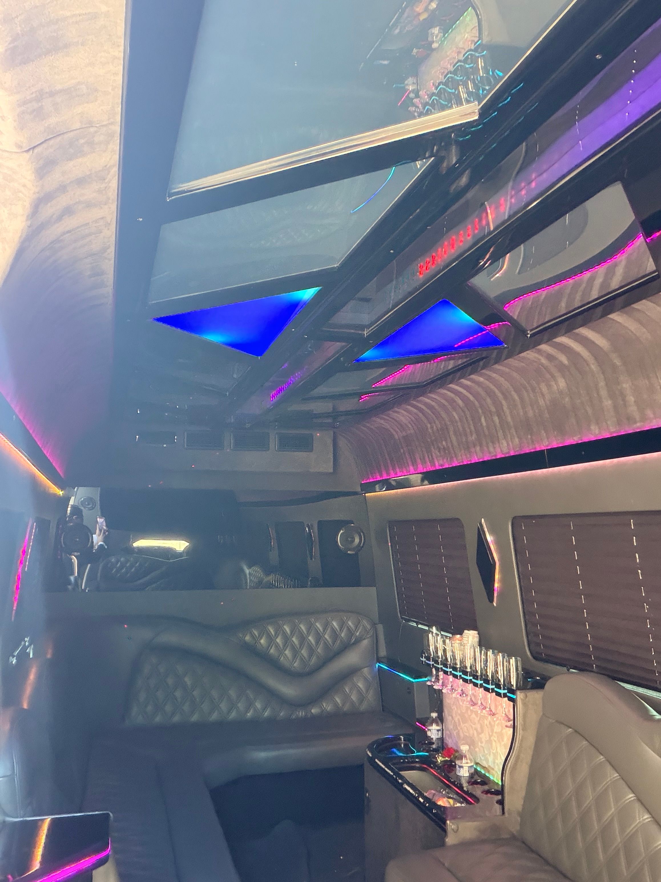 Luxury party limo interior with quilted black leather seats, triangular blue LED ceiling panels, purple neon accent lighting and a built-in bar stocked with champagne flutes