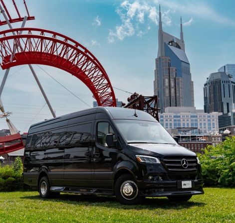 Sleek black passenger van parked on a grassy lawn in front of a red amusement-park arch with the Nashville skyline and twin-spired skyscraper under a blue sky.