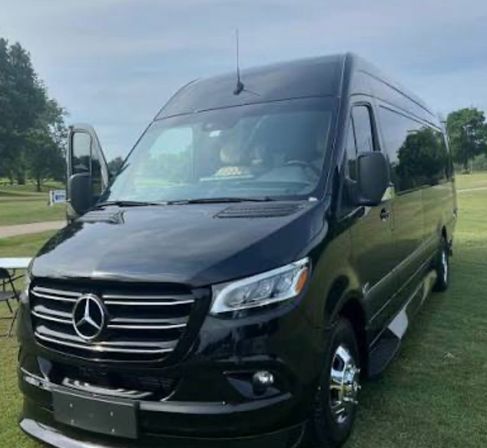 Black Mercedes‑Benz Sprinter passenger van parked on a grassy field, front three-quarter view with driver door ajar, chrome grille and hubcaps, trees and cloudy sky in background.