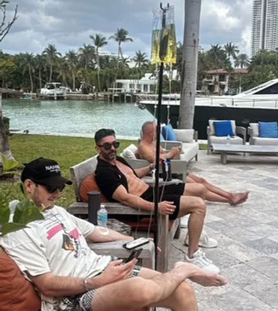 Three men lounging on a tropical waterfront patio beside a marina and luxury yacht, palm trees overhead and IV drip stands next to their lounge chairs.