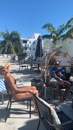 Two people lounging on a sunny rooftop patio with palm trees, a striped towel, rattan egg chair and umbrella against a clear blue sky and white buildings.