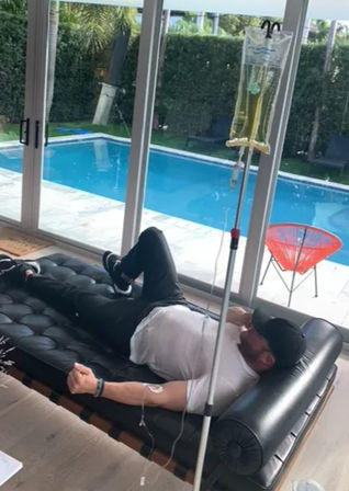 Person reclining on a black leather daybed in a living room, receiving an IV drip beside sliding glass doors overlooking a residential backyard swimming pool and patio chair.